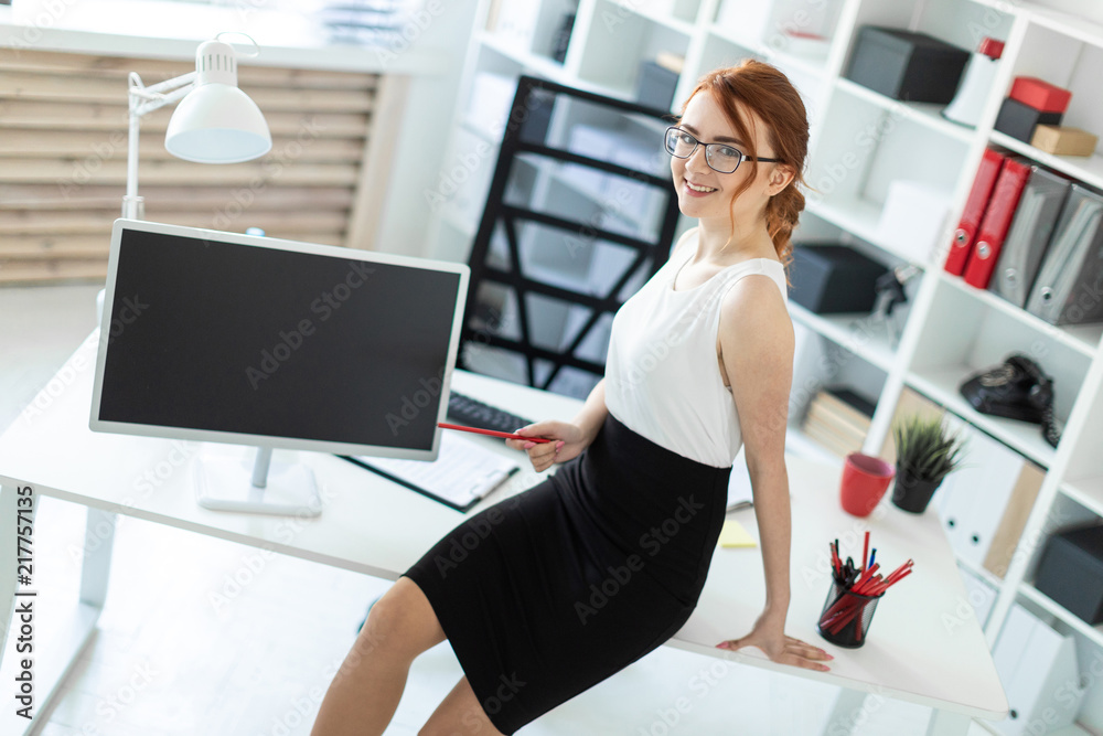 A beautiful young girl in the office sat down on the table and was holding a red pencil in her hand. Next to the girl is a monitor. A beautiful young girl in the office sat down on the table and was holding a red pencil in her hand. Next to the girl is a monitor.