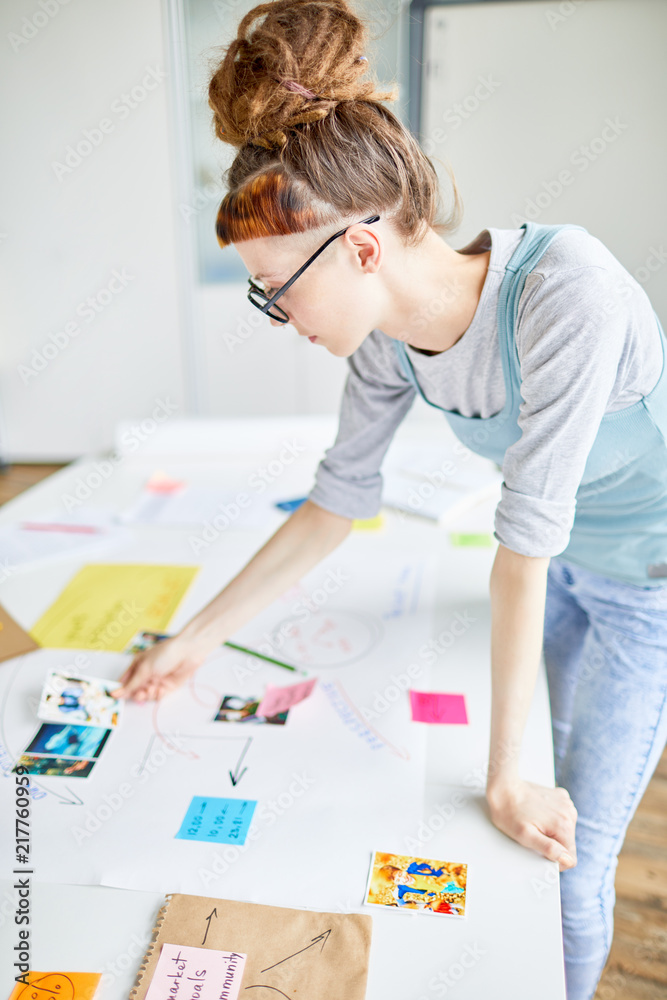 Teenage student standing by desk and looking through pictures while selecting some for her school project Teenage student standing by desk and looking through pictures while selecting some for her school project