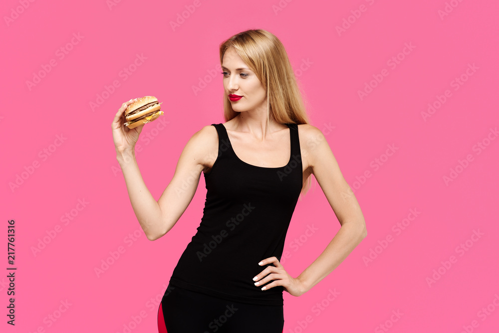 Young beautiful girl on a pink background holds a burger. The concept of an unhealthy diet Young beautiful girl on a pink background holds a burger. The concept of an unhealthy diet