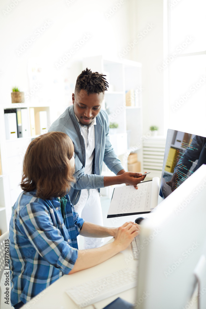 One of young it professionals explaining his colleague how to code data while pointing at computer monitor One of young it professionals explaining his colleague how to code data while pointing at computer monitor