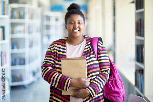 Positive confident overweight African-American student girl in colorful cardigan holding sketchpad and smiling at camera in library
