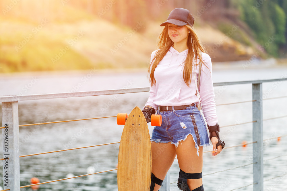 Obraz premium Young beautiful girl standing with longboard near the mountain lake beach