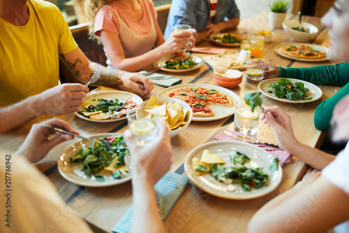 Hands of young people sitting around served table during dinner and enjoying salad