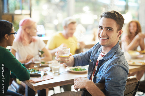 Young smiling man cheering up with drink while looking at you during dinner with friends