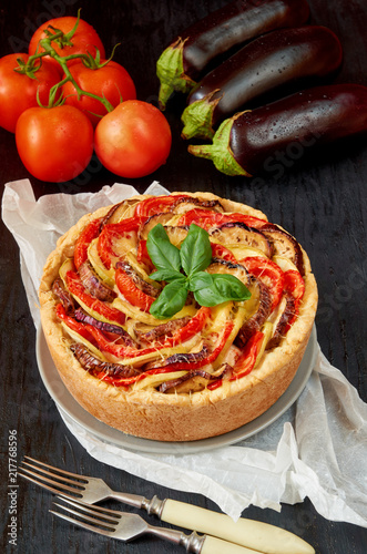 Pie with eggplants, tomatoes and cheese on the gray plate decorated with fresh basil leaves and vintage forks. Homemade vegetable tart on the black background. Side view