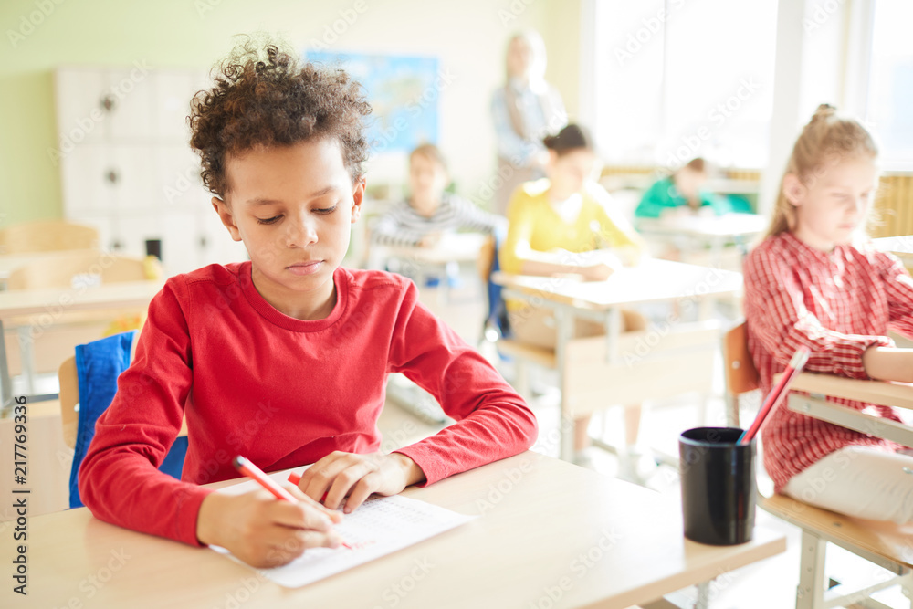 Serious schoolboy with Afro hairstyle sitting at table and marking answer in blank while passing exam in elementary school Serious schoolboy with Afro hairstyle sitting at table and marking answer in blank while passing exam in elementary school