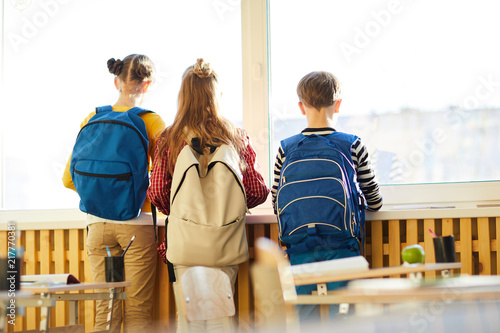 Rear view of dreamy school friends with satchels on backs looking out window while waiting for parents in classroom photo