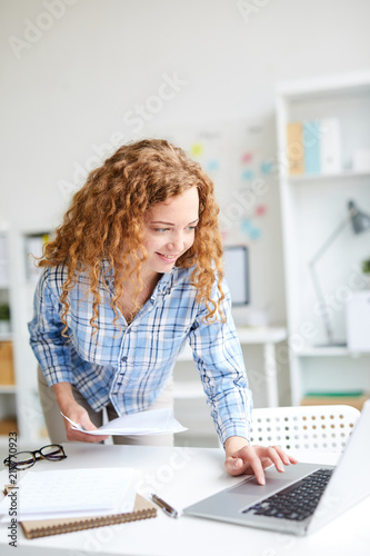 Young accountant with documents leaning over desk while looking for something in the net