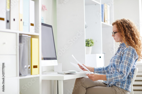 Businesswoman in casualwear reading papers while sitting by her desk in front of computer monitor