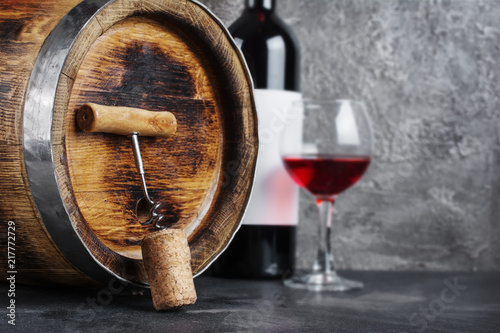 Red wine bottle with glass for tasting and wooden barrel with corkscrew in dark cellar