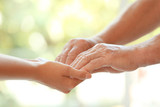 Young woman holding elderly man hands on blurred background, closeup. Help service