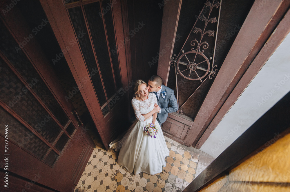 Beautiful newlyweds gently embrace near the ancient door and wall. A wedding portrait of a stylish groom and a cute bride in a lace dress in an old house.
