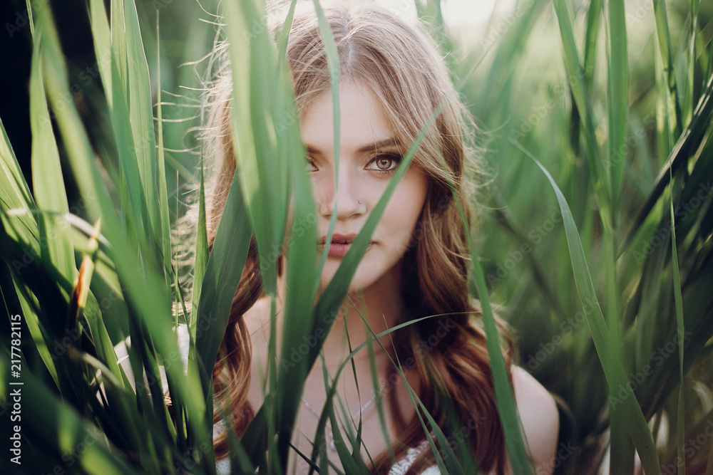 beautiful boho girl embracing in grass at sunset light near lake. attractive sensual young woman with amazing eyes and lips posing in white dress in green cane. save environment