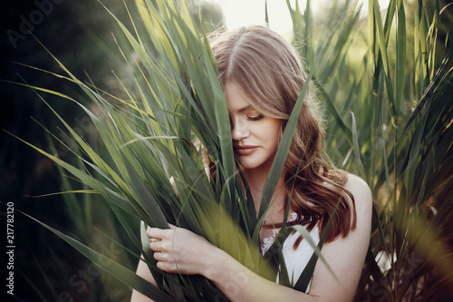 beautiful boho girl posing in grass at sunset light near lake. attractive sensual young woman embracing in cane near beach. summer vacation. save environment concept