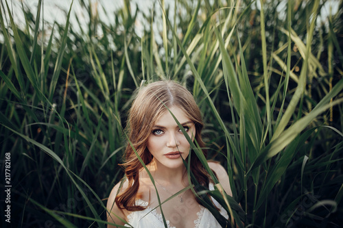 beautiful boho girl posing in grass at sunset light near lake. attractive sensual young woman embracing in cane near beach. summer vacation. save environment concept