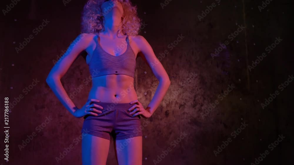 Slim woman with curly hair in shorts and sports bra breathing heavily after the workout in a dark gym. Tilt up real time medium shot Slim woman with curly hair in shorts and sports bra breathing heavily after the workout in a dark gym. Tilt up real time medium shot