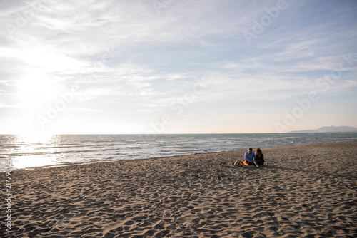 Young Couple Sitting On The Beach beside Campfire drinking beer