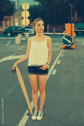 Woman in street vintage fashion wear standing on parking lot in front of road signs and looking away while holding longboard