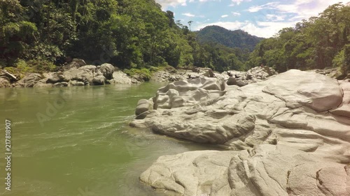 A wide shot of the calm green-hued Mulatos River winding through a rocky jungle valley near Tena in the Ecuadorian Amazon, lush forested hillsides and distant mountains, the camera panning left.