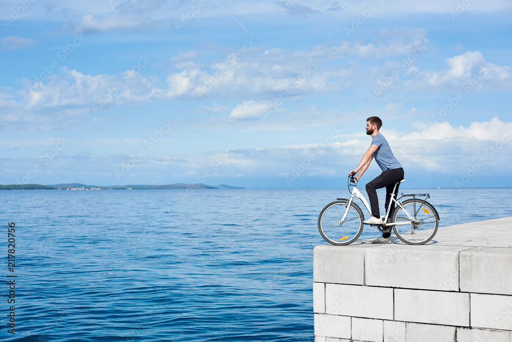 Fototapeta premium Young bearded man tourist on bicycle on high paved stone sidewalk enjoying clear blue sea water. Active holiday and tourism concept.