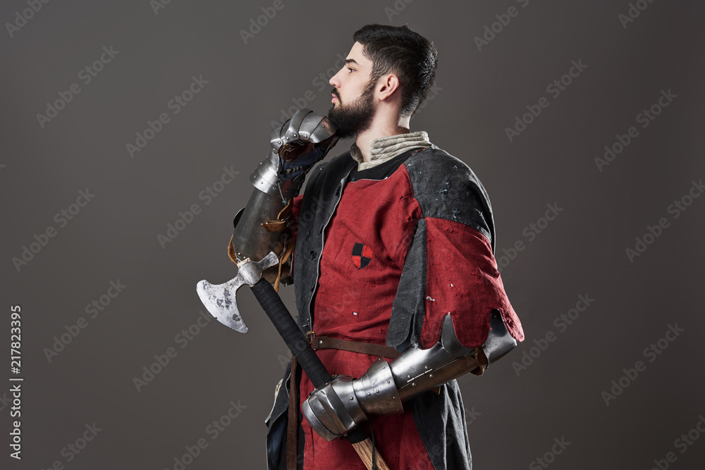 Medieval knight on grey background. Portrait of brutal dirty face warrior with chain mail armour red and black clothes and battle axe