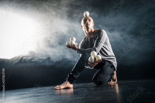 Tablou pe pânză Blond juggler sitting on the floor with white balls on black background