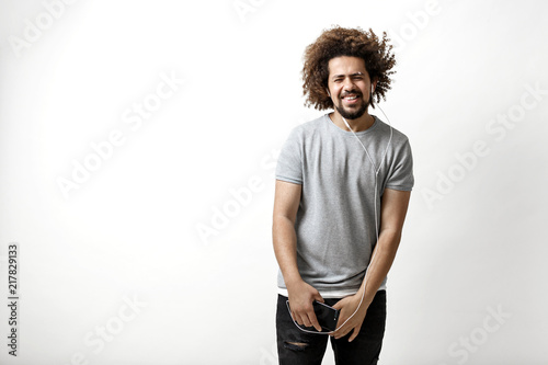 Slika na platnu A curly-headed handsome man wearing a gray T-shirt is standing with a joyous smile and listening to music in the earphones over the white background