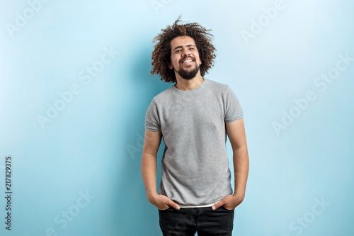 Fotografija A curly-headed handsome man wearing a gray T-shirt and ripped jeans is standing and broadly smiling with his hands in the pockets, over the blue background