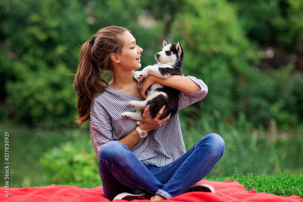 A beautiful smiling woman with a ponytail and wearing a striped shirt is cuddling with a sweet husky puppy while resting on the red blanket on the lawn. Love and care for pets. A beautiful smiling woman with a ponytail and wearing a striped shirt is cuddling with a sweet husky puppy while resting on the red blanket on the lawn. Love and care for pets.