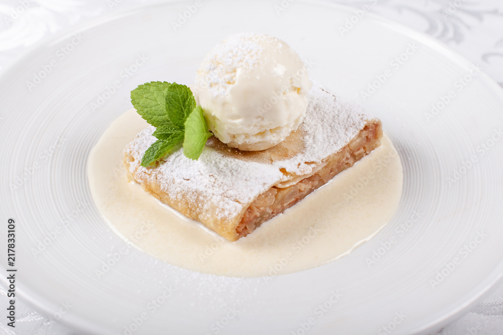 Fruit apple strudel cake served with ice cream, mint leaf and vanilla sauce. Classical austrian dessert on white plate. Sweet dessert in the restaurant. Morning light. Fruit apple strudel cake served with ice cream, mint leaf and vanilla sauce. Classical austrian dessert on white plate. Sweet dessert in the restaurant. Morning light.