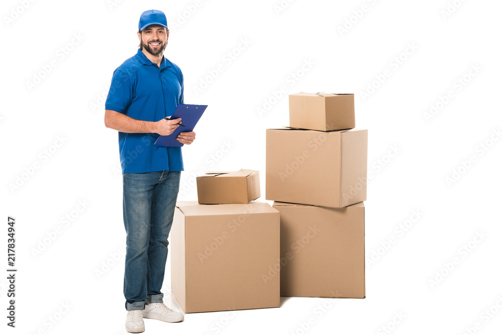 full length view of handsome young delivery worker holding clipboard and smiling at camera while standing near boxes isolated on white full length view of handsome young delivery worker holding clipboard and smiling at camera while standing near boxes isolated on white