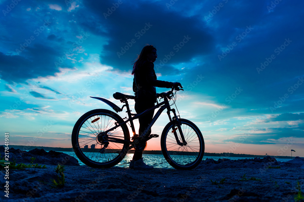 Fototapeta premium Silhouette of a young woman with a bicycle on the lake at sunset