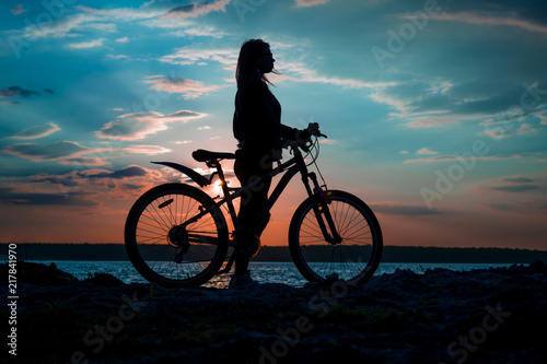 Silhouette of a young woman with a bicycle on the lake at sunset