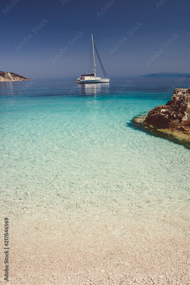 Beautiful calm azure blue lagoon with sailing catamaran yacht boat at anchor. Pure white pebble beach with rocks in the sea