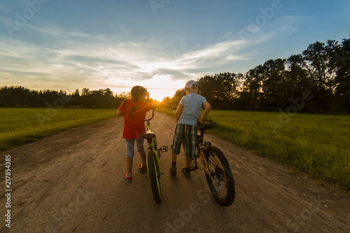 childs riding one bike together on sunny summer evening. sitting on bicycle rack. Family of two people enjoying traveling in scenic field over sunset sky background.