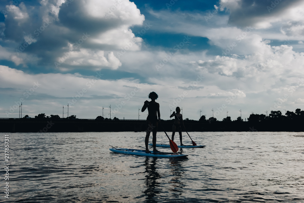 silhouettes of athletic women paddle boarding on river with cloudy sky