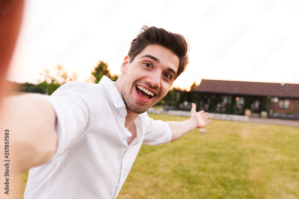 Image of joyful european man wearing white shirt laughing, while taking selfie during walk on nature near country house Image of joyful european man wearing white shirt laughing, while taking selfie during walk on nature near country house