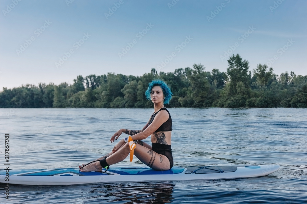 attractive tattooed sportswoman with blue hair relaxing on sup board on river