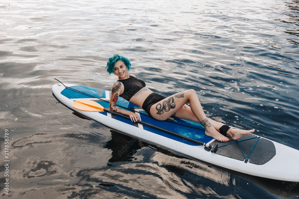 Naklejka premium attractive tattooed girl with blue hair lying on paddle board on river