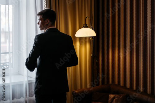 A man in a business suit is standing by the window in the hotel room.The groom in wedding day