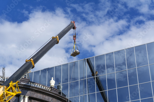 telescopic boom of a construction crane lifts the load against the mirror wall of the building reflecting the sky photo