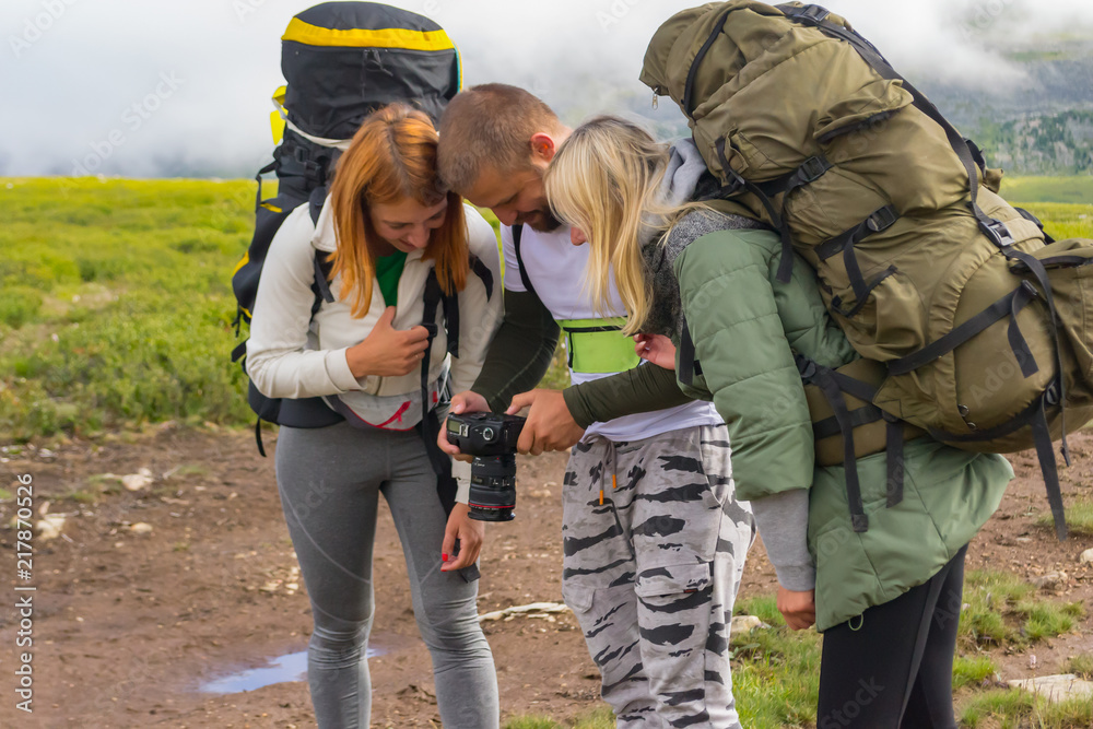 three young men, tourists, a guy and two girls on the sides with large backpacks for a walk in the Altai mountains, look at the photo from the camera, causing them funny emotions in the background fog