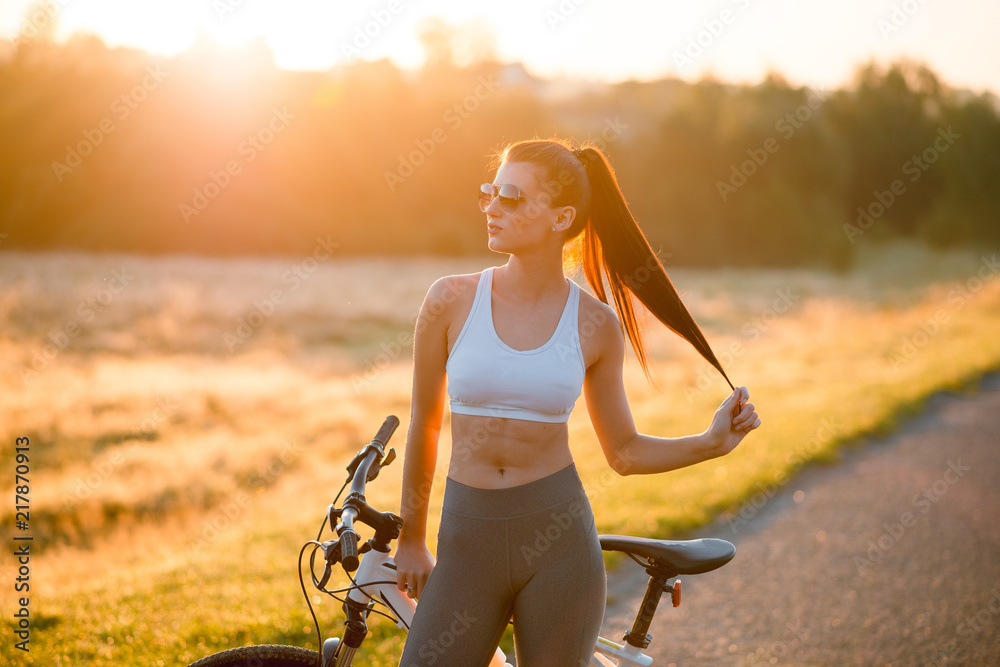 Close up portrait of attractive fit and sporty woman in sportswear standing with bicycle on the countryside road near the field on sunset. Close up portrait of attractive fit and sporty woman in sportswear standing with bicycle on the countryside road near the field on sunset.