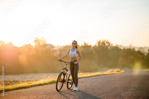 Fototapeta Naklejka Na Ścianę i Meble -  Beautiful happy fit and sporty woman in sportswear standing with bicycle on the countryside road near the field on sunset.