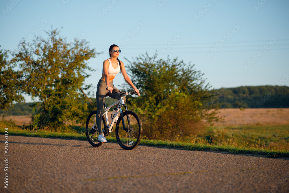 Beautiful young sporty brunette woman in sportswear riding bicycle outdoors on asphalt countryside road on sunny summer day.