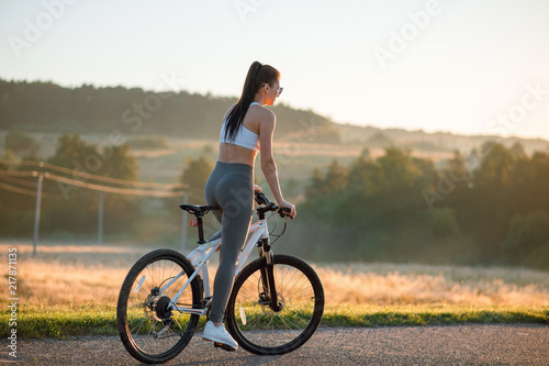 Back view shot of young sporty brunette woman in sportswear riding bicycle outdoors on asphalt countryside road on sunny summer day. © WellStock