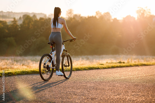 Rear view shot of young sporty brunette woman in sportswear riding bicycle outdoors on asphalt countryside road on sunset. Sun rays