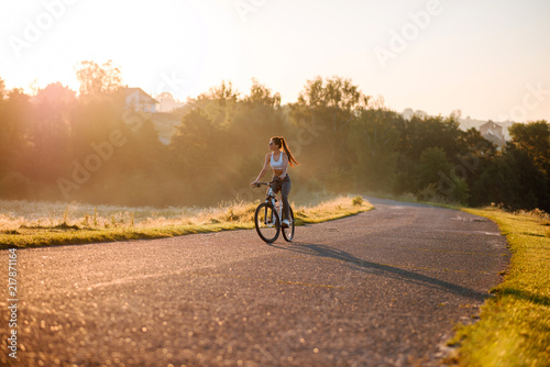 Young sporty brunette woman in stylish sportswear riding on bike on asphalt countryside road on sunset. Summer day © WellStock