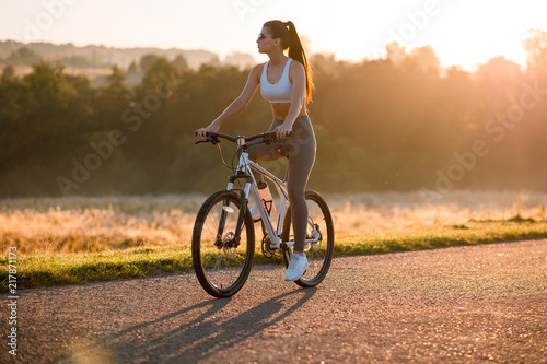 Beautiful young sporty brunette woman in stylish sportswear riding on bike on asphalt countryside road on sunset. Summer day