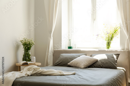 Blanket and pillows on grey bed in bright bedroom interior with flowers and window. Real photo © Photographee.eu